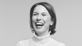 A black-and-white photograph of a smiling woman with short dark hair, wearing a light-coloured blouse and a ring on her finger. Her arms are crossed over her chest.