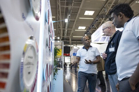 Ein Aussteller erklärt Besuchern die an einer Wand des Messestands ausgestellten Farbmuster.