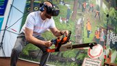 Mann mit VR-Brille simuliert Holzfällarbeit mit Motorsäge in einer interaktiven Messeinstallation vor Waldkulisse.