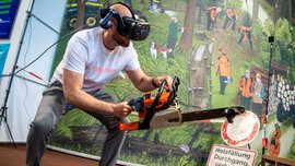 Mann mit VR-Brille simuliert Holzfällarbeit mit Motorsäge in einer interaktiven Messeinstallation vor Waldkulisse.