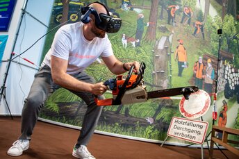 Mann mit VR-Brille simuliert Holzfällarbeit mit Motorsäge in einer interaktiven Messeinstallation vor Waldkulisse.