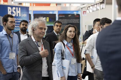 Eine Gruppe von Menschen nimmt an einer Messeführung teil und steht aufmerksam zuhöhrend an einem Messestand.