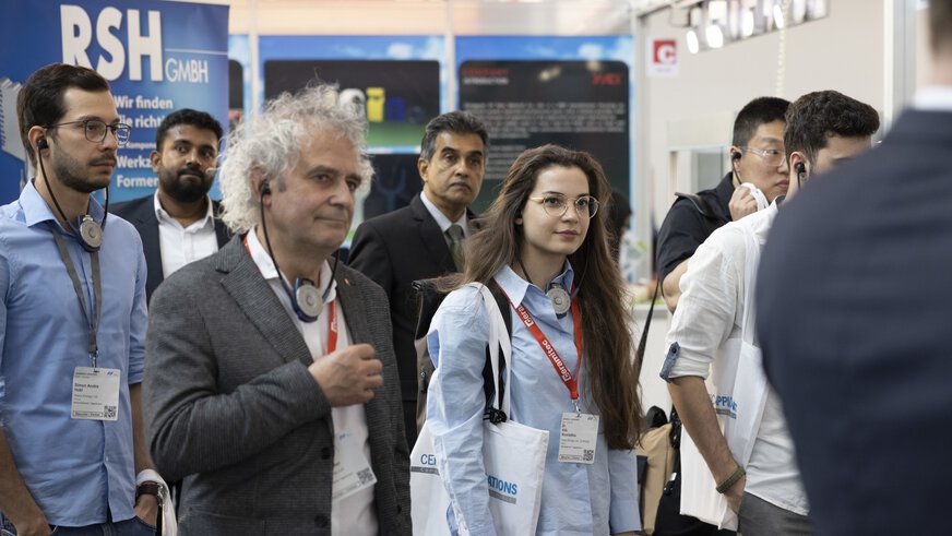 Eine Gruppe von Menschen nimmt an einer Messeführung teil und steht aufmerksam zuhöhrend an einem Messestand.