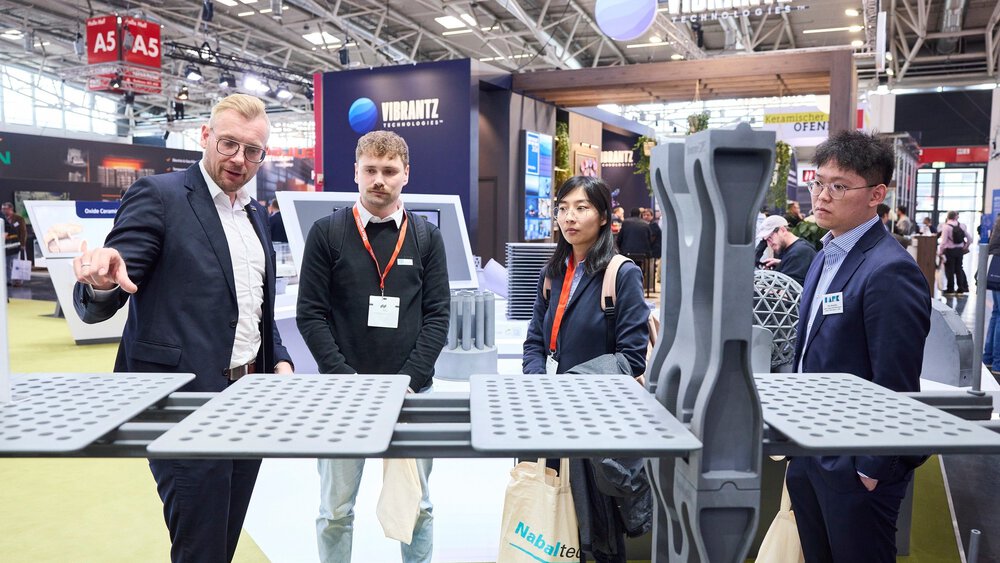 Vier Personen betrachten graue Platten mit Löchern, die an einem Stand auf der ceramitec präsentiert werden. Im Hintergrund sind weitere Stände zu sehen.