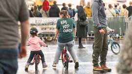 Eine Frau mit „Kids Velo Coach“-Shirt und ein kleines Mädchen sitzen je auf einem Fahrrad und fahren über einen abgetrennten Bereich auf der f.re.e.
