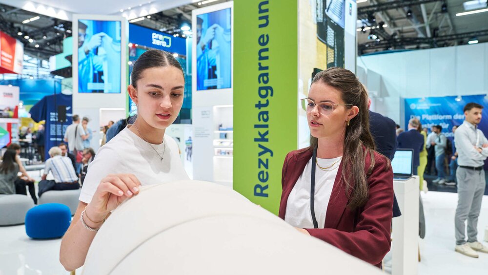 Two women examining a large white fabric element at a trade fair booth focused on recycled yarns and textiles.