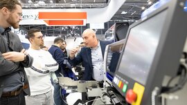 Zwei Männer lauschen interessiert einem Aussteller an einem Stand auf der productronica. Im Hintergrund sind weitere Personen zu sehen.