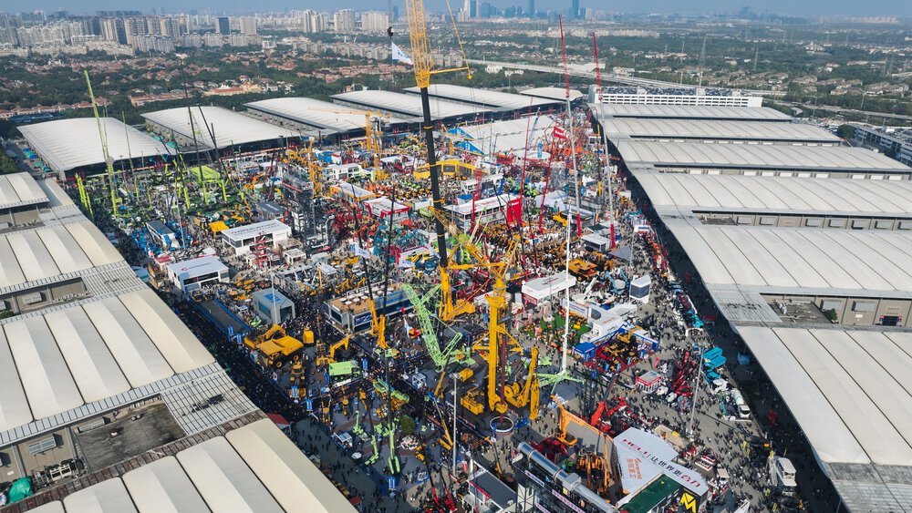 Aerial view of a large construction equipment exhibition featuring cranes and machinery, surrounded by crowds of people.
