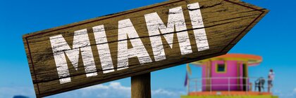 A wooden sign in the shape of an arrow with the word “MIAMI” printed on it against the blurred backdrop of a busy sandy beach.