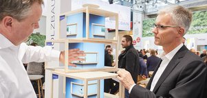 Zwei Männer in Businesskleidung begutachten das Holzmodell von mehreren übereinandergestapelten offenen Räumen an einem Stand auf der BAU.
