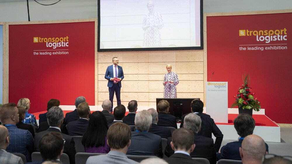 A man and a woman in business attire present to an interested audience on a stage at transport logistic.