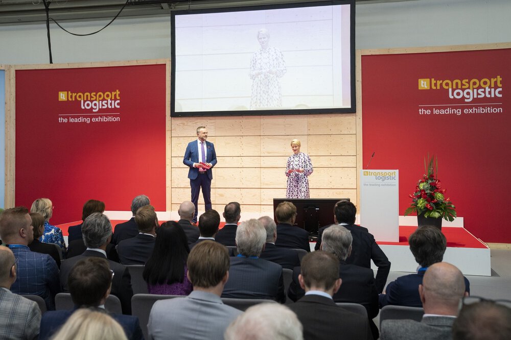 Ein Mann und eine Frau in Geschäftskleidung moderieren vor einem interessierten Publikum auf einer Bühne der transport logistic.