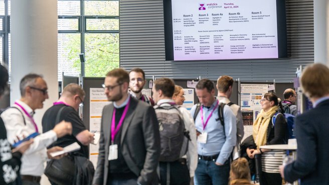 Eine Gruppe von Personen steht in einem Konferenzraum. An einer Wand hängt ein Bildschirm mit dem Konferenzprogramm