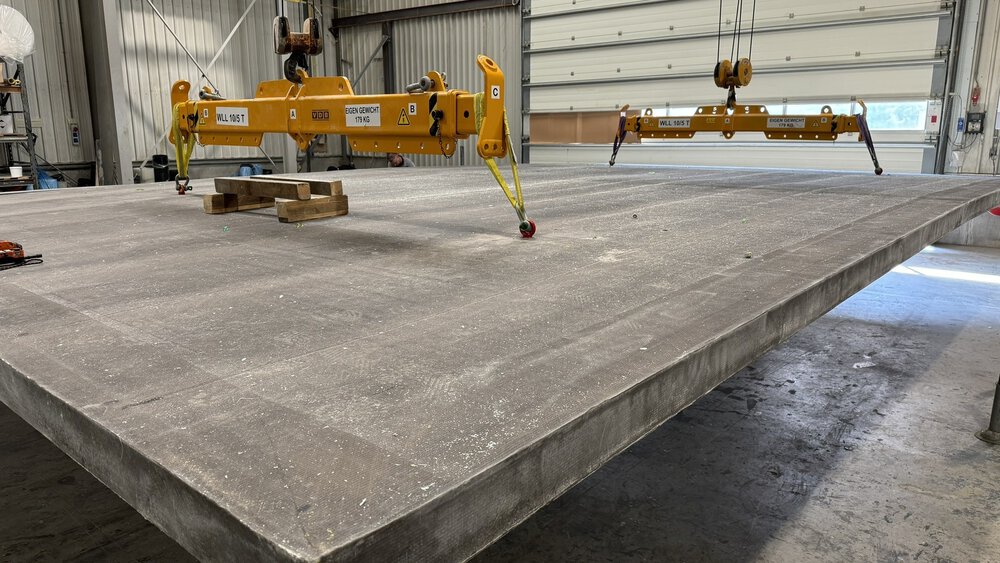 Große Betonplatte, die in einer Industriehalle an einer gelben Hebevorrichtung aufgehängt ist.