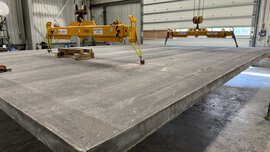 Große Betonplatte, die in einer Industriehalle an einer gelben Hebevorrichtung aufgehängt ist.