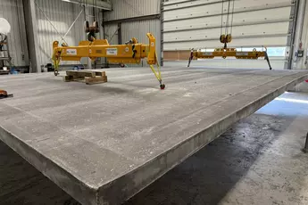 Large concrete slab suspended from a yellow lifting device in an industrial hall.
