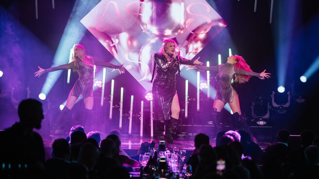 Ásdís appears on stage with two dancers, spotlights illuminate the scene while a rapt audience watches in the foreground.