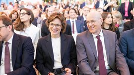 Close-up of smiling guests of honor in the front row of a conference, with the audience in the background.