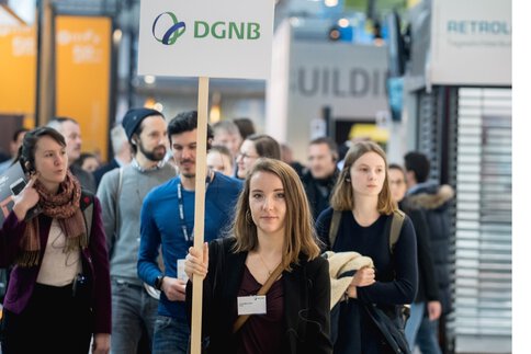 Eine Frau mit einem Schild, auf dem das „DGNB“-Logo zu sehen ist, führt eine Gruppe Besucher über die BAU.