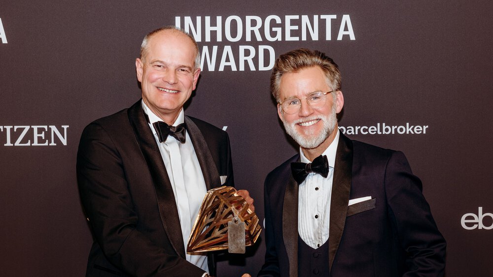 Two smiling men in suits shake hands in front of a photo wall of the INHORGENTA AWARD and hold a trophy.