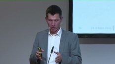 Ein Mann im grauen Anzug spricht in ein Mikrofon vor einer Leinwand.