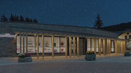 Modernes Gebäude mit Glasfassade und warmem Licht in der Nacht, umgeben von Bäumen und einem Sternenhimmel.