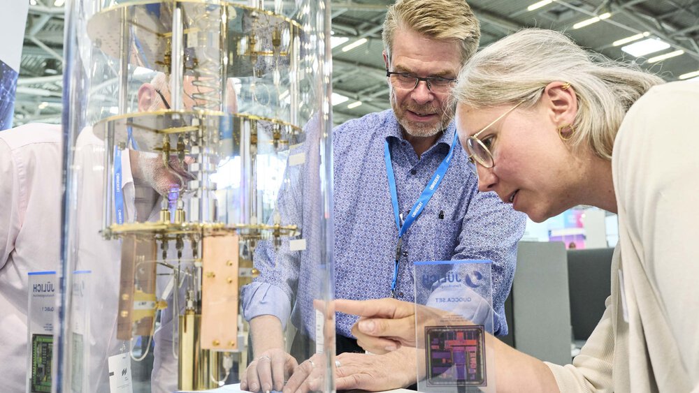 Three people look with interest at a quantum computer model at a technology fair, surrounded by information boards and exhibits.