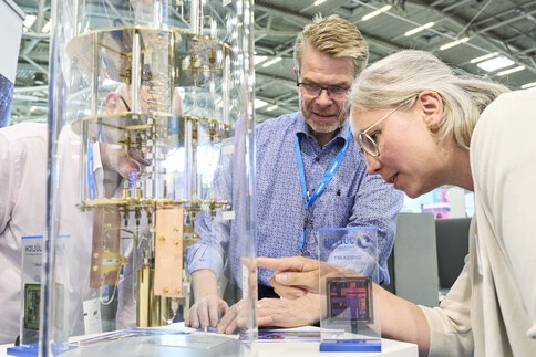 Drei Personen betrachten interessiert ein Quantencomputermodell auf einer Technikmesse, umgeben von Infotafeln und Exponaten.