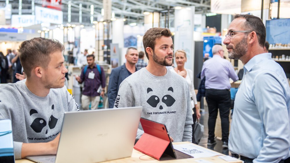 Zwei junge Männer in grauen Sweatshirts mit einem Panda‑Logo und der Aufschrift „The Fortunate Planet“ stehen an einem Messestand mit Laptops und sprechen mit einem Besucher; im Hintergrund sind weitere Messegäste und Stände in einer großen Halle zu sehen.