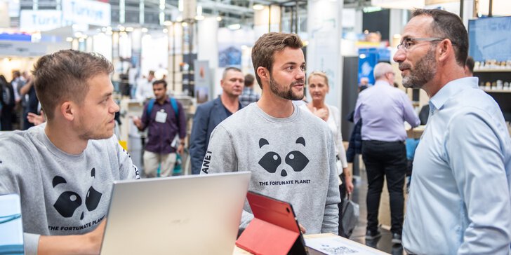 Zwei junge Männer in grauen Sweatshirts mit einem Panda‑Logo und der Aufschrift „The Fortunate Planet“ stehen an einem Messestand mit Laptops und sprechen mit einem Besucher; im Hintergrund sind weitere Messegäste und Stände in einer großen Halle zu sehen.