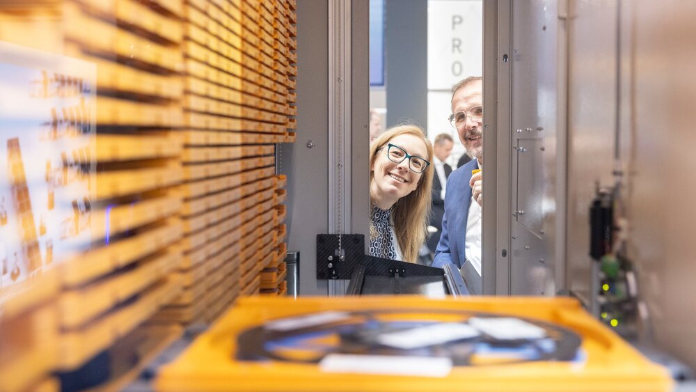 Ein Mann und eine Frau mit Brille blicken lächelnd in das Innere einer Maschine aus Metall, die mit gelben Elementen bestückt ist.
