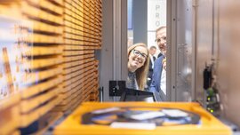 Ein Mann und eine Frau mit Brille blicken lächelnd in das Innere einer Maschine aus Metall, die mit gelben Elementen bestückt ist.