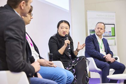 Four people sit in armchairs and talk to each other. One of the people is holding a microphone and gesticulating while talking