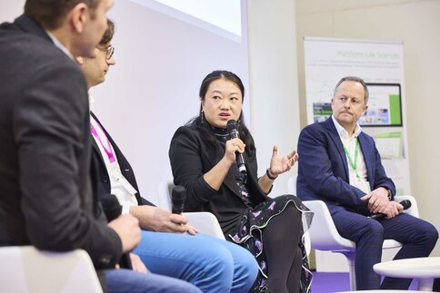Four people sit in armchairs and talk to each other. One of the people is holding a microphone and gesticulating while talking