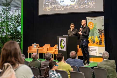 Two men standing on a stage, one speaking into a microphone in front of a seated audience.