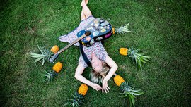 Eine lächelnde Frau mit hellen Haaren und in einem geblümten Sommerkleid liegt mit einer bunten Gitarre auf einer grünen Wiese und ist umringt von Ananasfrüchten.