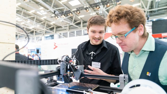 Zwei Personen inspizieren einen ausgestellten Roboter, der Elemente auf einer Leiterplatte platziert.