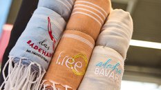 Drei zusammengerollte, bestickte Handtücher mit den Aufschriften „das Badehaus“, „f.re.e“ und „aloha BAVARIA“ in Nahaufnahme.