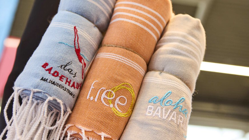 Drei zusammengerollte, bestickte Handtücher mit den Aufschriften „das Badehaus“, „f.re.e“ und „aloha BAVARIA“ in Nahaufnahme.