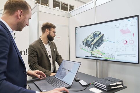 Zwei Männer stehen vor einem Laptop und einem großen Bildschirm, auf dem eine Architektursoftware angezeigt wird.
