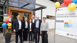 Eine Gruppe von sechs Personen steht vor einem Gebäude neben einem Schild, das die Feier zum 25-jährigen Bestehen des Bauzentrum Poing verkündet.