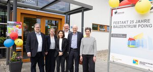 Eine Gruppe von sechs Personen steht vor einem Gebäude neben einem Schild, das die Feier zum 25-jährigen Bestehen des Bauzentrum Poing verkündet.