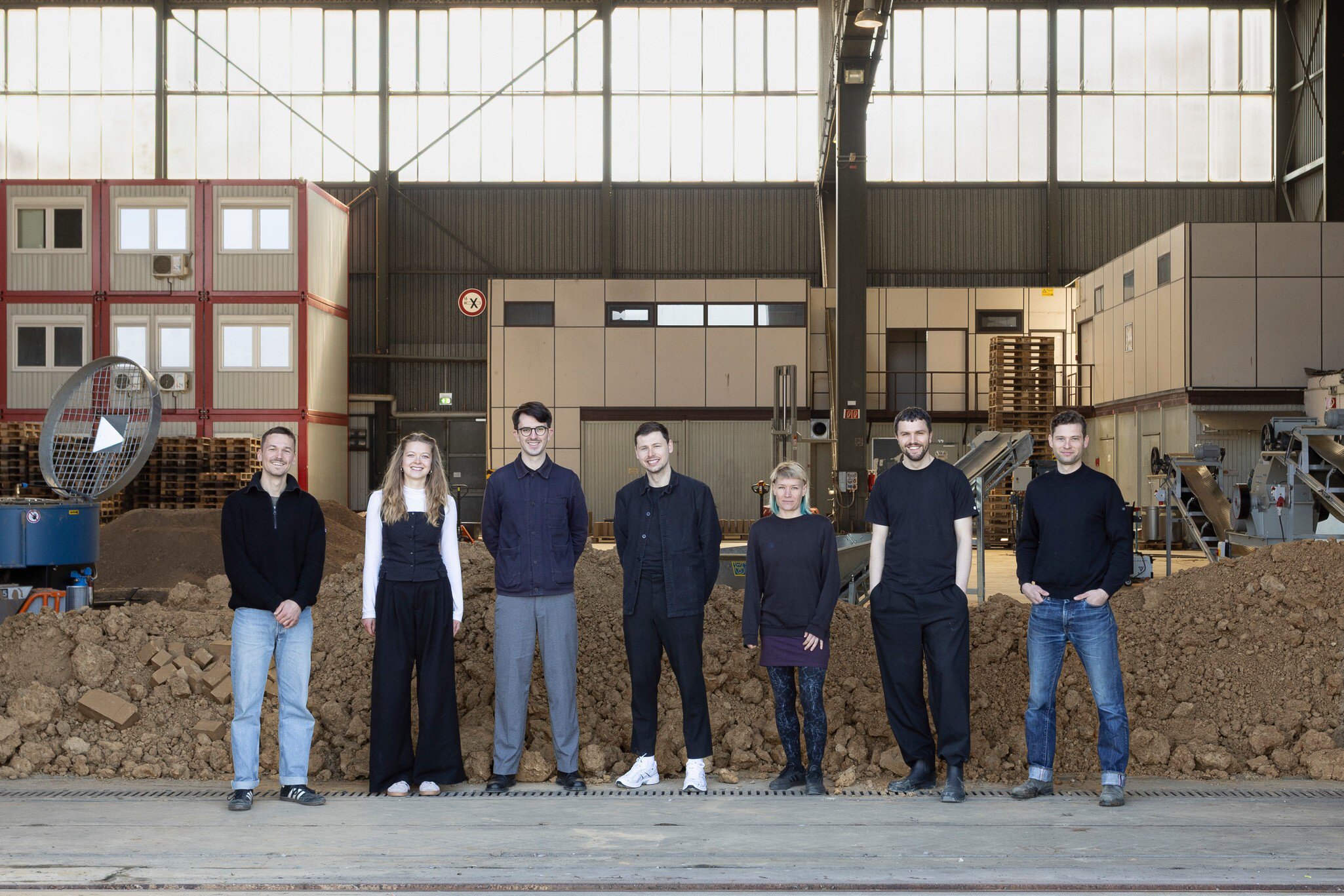 Seven people stand side by side in an industrial hall in front of a mound of earth, with machines and containers visible in the background.