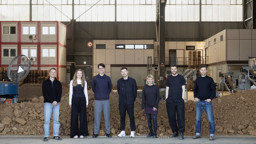 Seven people stand side by side in an industrial hall in front of a mound of earth, with machines and containers visible in the background.