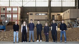 Seven people stand side by side in an industrial hall in front of a mound of earth, with machines and containers visible in the background.