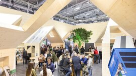 Messestand mit einer großen, hausähnlichen Holzkonstruktion, an deren Tischen zahlreiche Messebesucher sitzen, stehen und sich unterhalten.