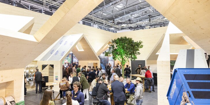 Messestand mit einer großen, hausähnlichen Holzkonstruktion, an deren Tischen zahlreiche Messebesucher sitzen, stehen und sich unterhalten.