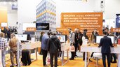People gather at a technology exhibition stand in orange design, with screens and information displays.