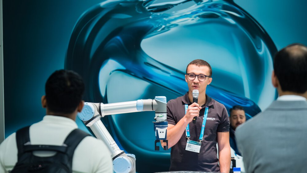 Ein Mann mit einem Mikrofon steht vor einer blauen Wand und hat einen silbernen Roboterarm vor sich stehen. Er spricht zu zwei Messebesuchern.