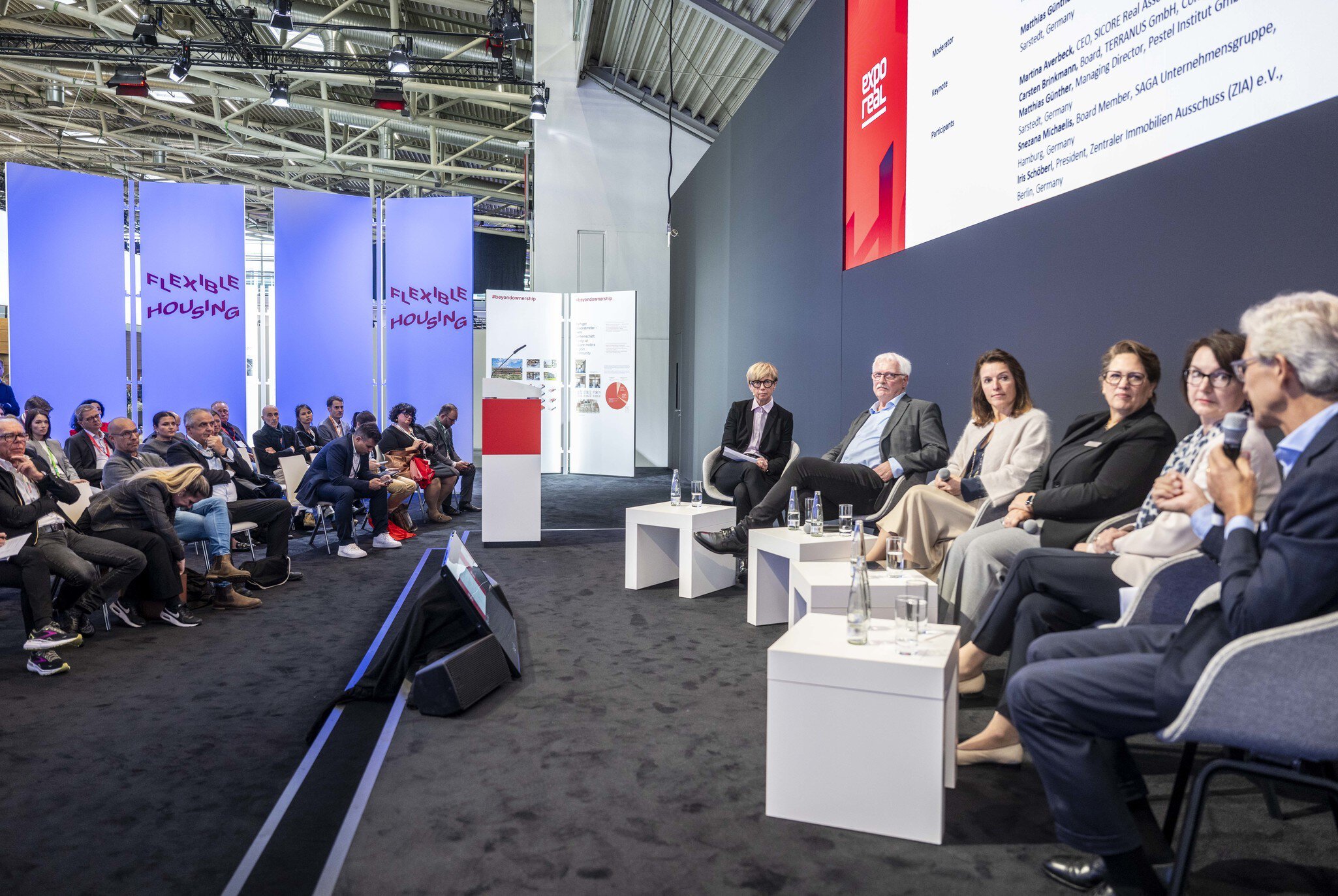 A panel discussion at EXPO REAL: Seven speakers are seated on stage in a row, addressing a large audience. The audience sits in several rows on the left, while the panelists on the right hold microphones with water glasses placed on small tables. Large blue banners reading “Flexible Housing” stand behind the audience.
